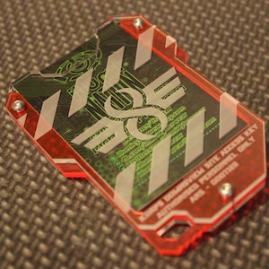 Op de afbeelding: Een rode plastic sleutelkaart met witte diagonale strepen en een zwart en groen ontwerp. De kaart heeft de tekst "ALPHA DOMINATED BYTE ACCESS KEY AUTHORISED PERSONNEL ONLY" en een websiteadres.