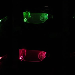 May include: Two pairs of glowing green and pink glasses with a logo on the lenses. The glasses are made of clear plastic and have a futuristic design.