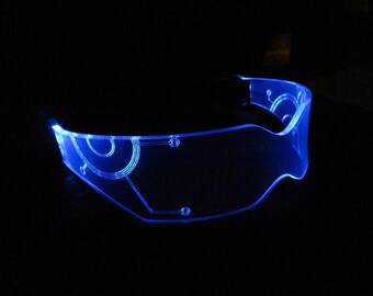 May include: A pair of blue, futuristic-style glasses with a clear plastic frame and a circuit board design. The glasses are lit up with blue LED lights.