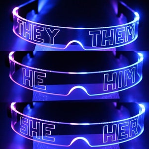 Könnte beinhalten: Drei Paar transparente LED-Brillen aus Kunststoff mit verschiedenen Pronomen darauf. Das obere Paar sagt "They Them", das mittlere Paar sagt "He Him" und das untere Paar sagt "She Her".