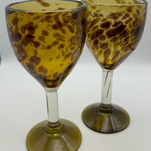 May include: Two amber-colored glass goblets featuring a speckled brown pattern throughout the bowl. Each goblet has a clear glass stem and a wide, circular amber base. The glass has a smooth, rounded rim and a distinct, hand-blown appearance.