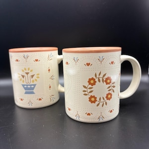 May include: Two off-white ceramic mugs with a peach-colored rim. The mugs feature a repeating grid pattern with small dots, orange hearts, and blue floral accents. One mug displays a central floral wreath, while the other shows a stylized flower pot.