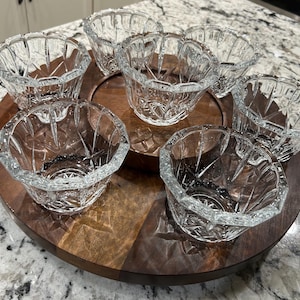 May include: A set of seven clear glass serving bowls with a starburst design, arranged on a two-toned wooden serving tray. The bowls have a faceted, scalloped rim and are ideal for serving dips, snacks, or small portions.