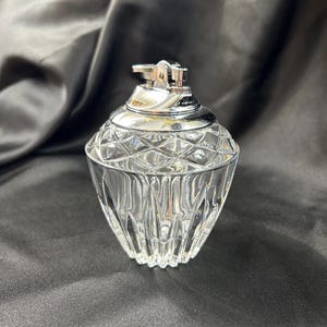 May include: A clear glass lighter with a silver metal top. The glass base has a diamond pattern and vertical lines. The lighter is on a dark background.