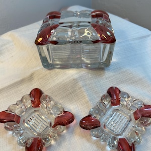 May include: A clear glass trinket box with a red trim and two matching ashtrays. The box is rectangular with a lid, and the ashtrays are square with a decorative red design. The glass has a ribbed texture.