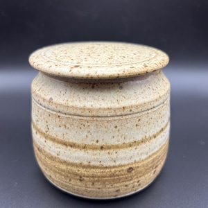 May include: A cylindrical ceramic jar with a matching lid, featuring a speckled beige and tan glaze. The body of the jar displays horizontal bands of darker brown tones, with a textured, earthy finish throughout the entire exterior surface.