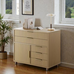 May include: A light beige wooden dresser with three drawers and two doors. The dresser has vertical ribbed detailing on the front and gold-colored handles. A small lamp and books sit on top of the dresser.