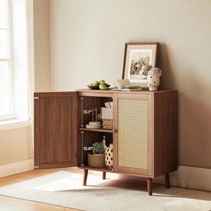 May include: A brown wooden cabinet with two doors, one open. The cabinet has a woven cane door and two shelves. The open door reveals 13.1" and 15.8" of interior space. The cabinet is on short, angled legs. A framed picture and decor sit on top.