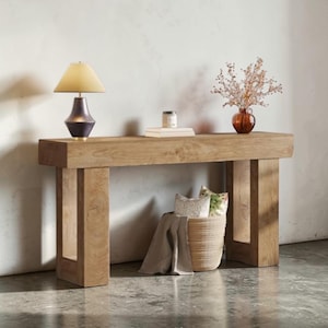 May include: A light brown wooden console table with a rectangular top and two supporting legs. A lamp with a beige shade and dark base sits on the table, along with books, a vase with dried flowers, and a woven basket with pillows and a blanket.