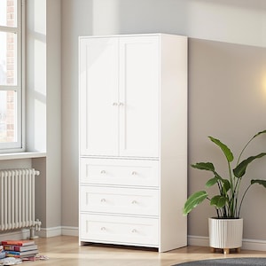 May include: A tall, white wooden wardrobe with two upper doors and three drawers. The doors and drawers have silver-toned handles. The wardrobe is placed against a light gray wall, with a window and radiator visible on the left. A potted plant is on the right.