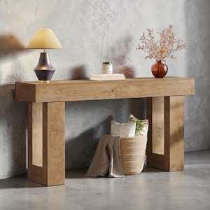 May include: A light brown wooden console table with a lamp, books, and a vase of flowers on top. A woven basket with pillows and a blanket sits below. The lamp has a gold shade and a dark base.