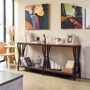 May include: A long rectangular console table with a dark brown wooden top and a black metal frame featuring geometric supports. The table has two levels, with the lower shelf holding books, a gray vase, white animal figurines, and a woven basket.