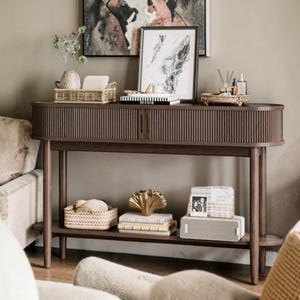 May include: A dark brown wooden console table with a rounded, fluted front design and two sliding doors. The table features a lower shelf for storage and four slender legs. It is topped with various decorative items, including a woven basket.