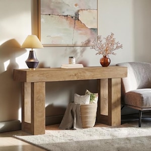 May include: A rectangular light brown wooden console table with thick block legs. On top sits a dark purple lamp with a cream shade, a small white box, and a brown glass vase with white floral branches. A woven basket with fabric sits underneath.