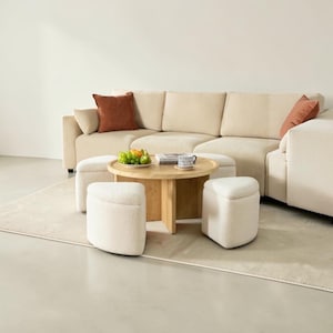 May include: A sectional sofa with cream-colored corduroy upholstery and rust-colored throw pillows. A round wooden coffee table holds a plate of fruit and a mug. Two small, off-white, upholstered ottomans are in front of the table, and a rug is underneath.