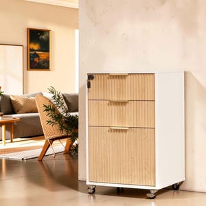 May include: A white mobile storage cabinet with three drawers featuring light wood-toned, vertically ribbed fronts. Each drawer has a slim, horizontal gold-colored handle. A small silver key hangs from a lock on the top drawer. The unit sits on wheels.