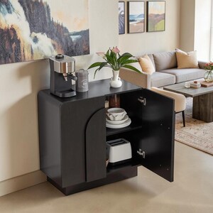 May include: A black wooden cabinet with rounded edges and a single hinged door. The top surface holds a silver and black espresso machine, a gray canister, and a white vase with green leaves. Inside, white bowls and a white toaster are visible.