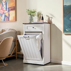 May include: A white wooden tilt-out trash cabinet featuring a single top drawer with a black knob and a lower compartment with a black handle. The cabinet has a dark brown top surface and a vertical plank design on the front panel of the tilt-out door.