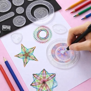 Könnte beinhalten: Eine Sammlung klarer Kunststoff-Spirograph-Zeichenwerkzeuge und Buntstifte auf einer weißen Oberfläche. Die Werkzeuge werden verwendet, um komplizierte geometrische Designs mit farbigen Stiften zu erstellen. Eine Hand zeichnet mit einem Stift ein Design.