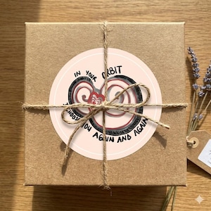 May include: A small, square, brown cardboard box tied with twine. A round sticker on top reads "In Your Orbit, I Choose You Again and Again" in black text. The sticker has a pink background with a heart design.