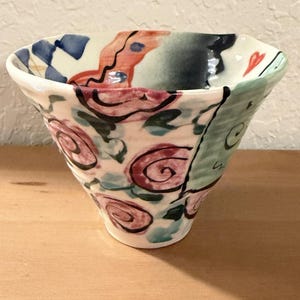 May include: A hand-painted ceramic bowl with a flared shape. The bowl features a cream-coloured base adorned with red, pink, and black floral designs. The rim has blue, red, and green accents. The bowl sits on a wooden surface.
