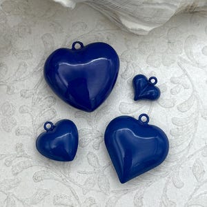 Op de afbeelding: Vier glanzende, marineblauwe hartvormige bedels in verschillende maten. Elke bedel heeft een kleine lus aan de bovenkant om aan sieraden of andere knutselwerken te bevestigen. De bedels zijn gerangschikt op een getextureerd, crèmekleurig oppervlak met schelpenaccenten.