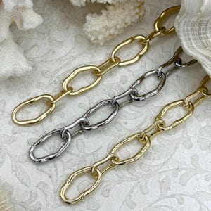 May include: Three chain necklaces in gold and silver tones. The chains have an oval link design. The necklaces are displayed on a white surface with coral and seashells in the background.