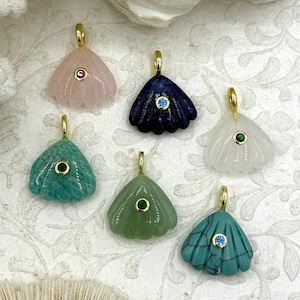 May include: Six gemstone pendants in various colors, including pink, blue, green, and white. Each pendant has a gold-colored bail and a small gemstone accent. The pendants are arranged on a patterned surface, with a seashell in the background.