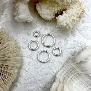 May include: A collection of silver-colored jump rings in various sizes, arranged on a white, patterned surface. The rings are open, ready for use in jewelry making or crafts. The background includes white coral and other natural elements.
