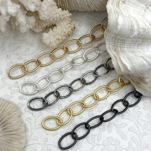 May include: Close-up of five different metal chain styles. The chains are gold, silver, black, and a matte gold color. The chains are oval-linked and are displayed on a white patterned surface.