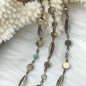 Può includere: Primo piano di tre fili di collane di perline. Le collane presentano una varietà di perline nelle tonalità dell'oro, del viola iridescente e del trasparente. Le perline sono collegate da delicate catene dall'aspetto antico. Lo sfondo è una superficie morbida, bianca e strutturata.