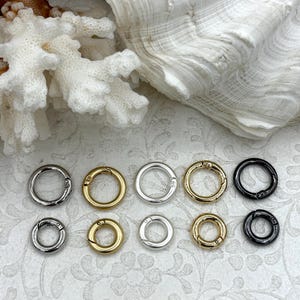 May include: An assortment of spring ring clasps in various metallic finishes, including silver, gold, and black. The clasps are arranged in two rows, showcasing their circular design and secure closure mechanism. The background features white coral and a seashell.