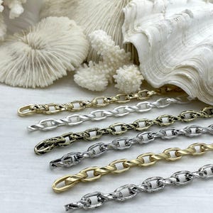 May include: Close-up of six metal chain necklaces in various finishes: gold, silver, and antique bronze. The chains have an oval link design. The background features white coral and a seashell, suggesting a coastal theme.