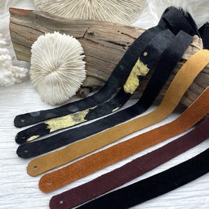 May include: A collection of leather straps in various colors, including black with gold accents, tan, brown, and burgundy. Each strap has a small hole at one end. The straps are displayed on a piece of driftwood.