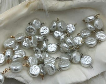 May include: A collection of lustrous, white, coin-shaped pearls with gold-colored connectors. The pearls are arranged in strands, resting inside a white, shell-shaped dish, against a backdrop of white, fluffy material.