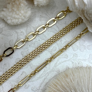 May include: Three gold-colored chain necklaces with different link styles. The top chain has large oval links, the middle chain has a textured, woven appearance, and the bottom chain features rectangular links. The chains are displayed on a white surface with coral.