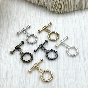 May include: A collection of toggle clasps in various metallic finishes, including silver, gold, and black. Each clasp features a ring and a bar, designed for jewelry making. The clasps are arranged on a textured white surface, showcasing their different colors and styles.