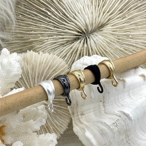 May include: A collection of five metal ear cuff earrings in silver, black, gold, and black, displayed on a wooden dowel. The earrings have a curved design with a hook. The background features a seashell and coral.