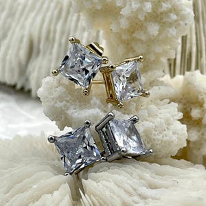 May include: Two pairs of square-cut stud earrings. One pair is gold-toned with clear, square-cut stones. The other pair is silver-toned with similar clear, square-cut stones. The earrings are displayed on a white, textured surface.