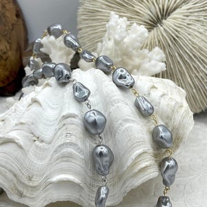 May include: A gray pearl necklace with irregular, baroque-style beads. The necklace is strung on a gold-colored chain and is displayed on a white seashell. The background includes white coral and a wooden element.