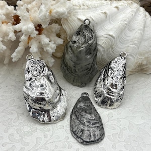 May include: Four silver-toned oyster shell pendants with a textured surface and a small loop at the top for hanging. The pendants are displayed on a white floral patterned surface with coral and a large shell in the background.