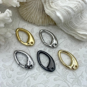 May include: An assortment of oval-shaped metal clasps in gold, silver, and black. Each clasp has a curved design with a small hole at the base. The clasps are arranged on a white surface with a floral pattern, alongside white coral and shell accents.