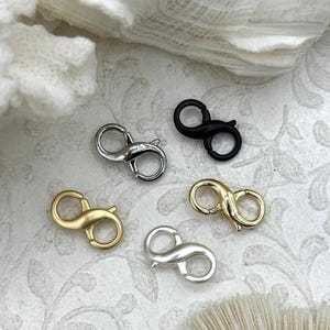 May include: An assortment of S-shaped clasps in various metallic finishes: gold, silver, and black. The clasps are displayed on a textured, off-white surface, showcasing their different colors and designs. Ideal for jewelry making.