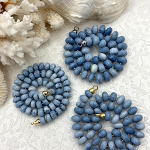 May include: Three necklaces made of light blue oval beads. Each necklace is arranged in a circular pattern, with gold or silver clasps. The necklaces are displayed on a white surface with a seashell and coral in the background.