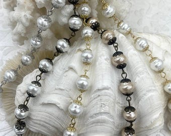 May include: Several strands of faux pearl beads in various colors, including white, cream, and champagne. The beads are strung together with metal chains in silver, gold, and bronze tones. The background features a white coral and seashell.