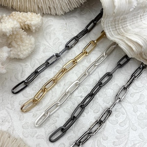May include: Close-up of five metal chain necklaces in various colors: black, gold, silver, dark gray, and distressed silver. The chains have an elongated oval link design. The background features white coral and a seashell.