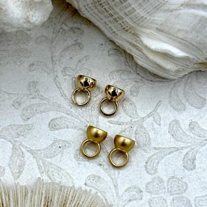 May include: Four small, gold-coloured jewellery findings with a half-dome shape and a circular loop. The findings are arranged on a white surface with a floral pattern and a piece of white coral.