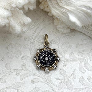 Könnte beinhalten: Runder Anhänger mit einem Bienenmotiv. Der Anhänger hat eine schwarze Mitte mit einer detaillierten Bienenillustration, umgeben von einem gold- und silberfarbenen Rahmen. Er hängt an einer kleinen, goldfarbenen Schlaufe.