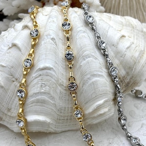 May include: Close-up of three delicate necklaces. One is gold-toned, one is silver-toned, and the other is gold-toned. Each necklace features a series of clear, round gemstones set in a linked chain design. The necklaces are displayed on a white, textured surface.