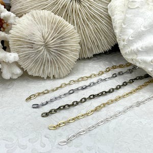May include: Close-up of five metal chain strands in gold, silver, and bronze tones. The chains are displayed against a backdrop of white coral and seashells. The chains are oval-linked and suitable for jewelry making.